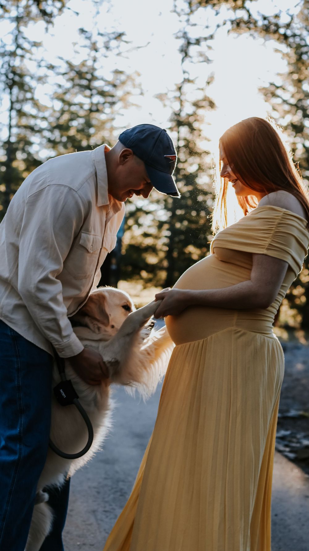 Pregnant woman in yellow dress and man in cap interact with a fluffy dog outdoors among sunlit trees.