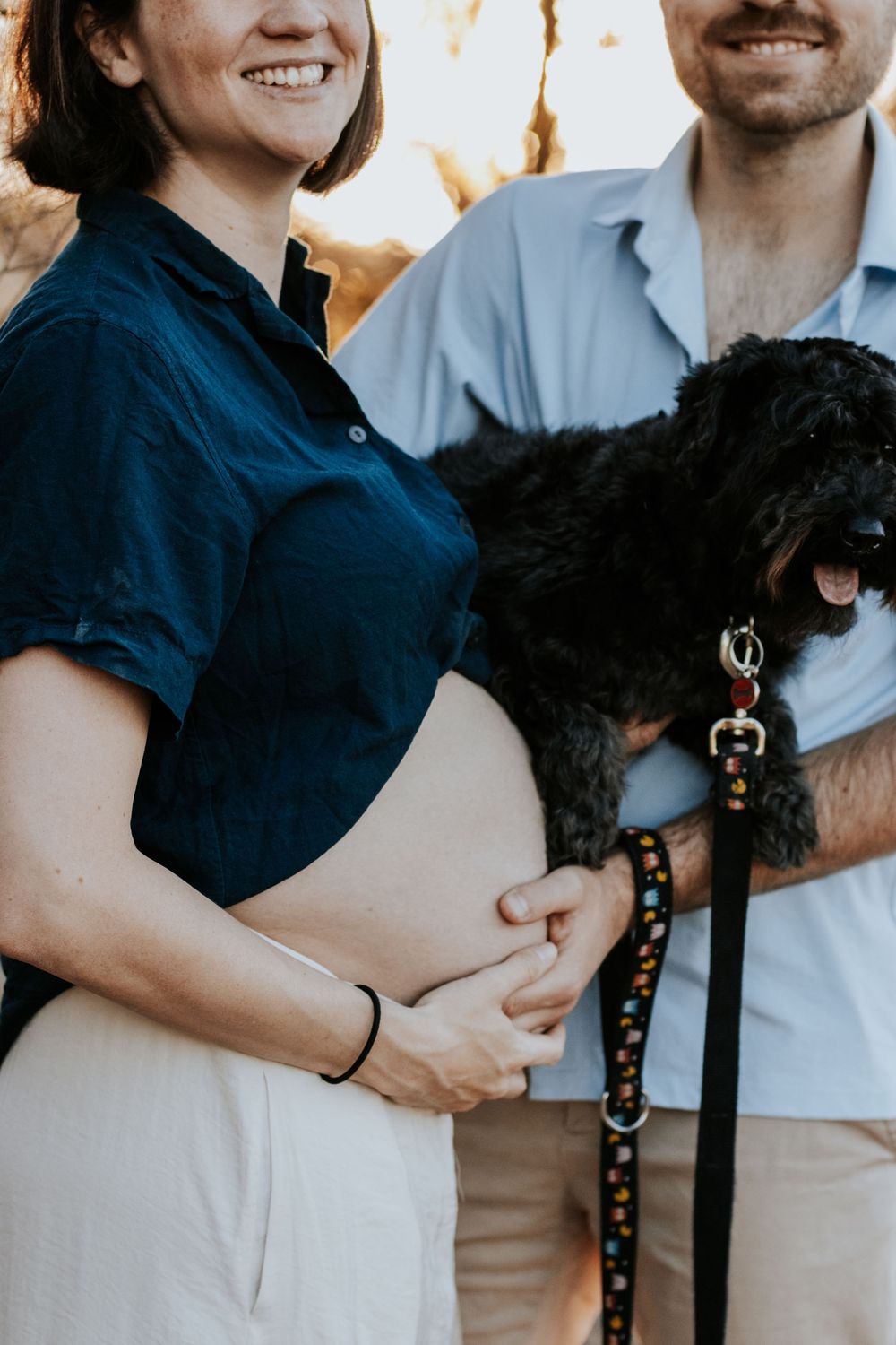 Pregnant woman in navy shirt with partner holding black dog on colorful leash outdoors.