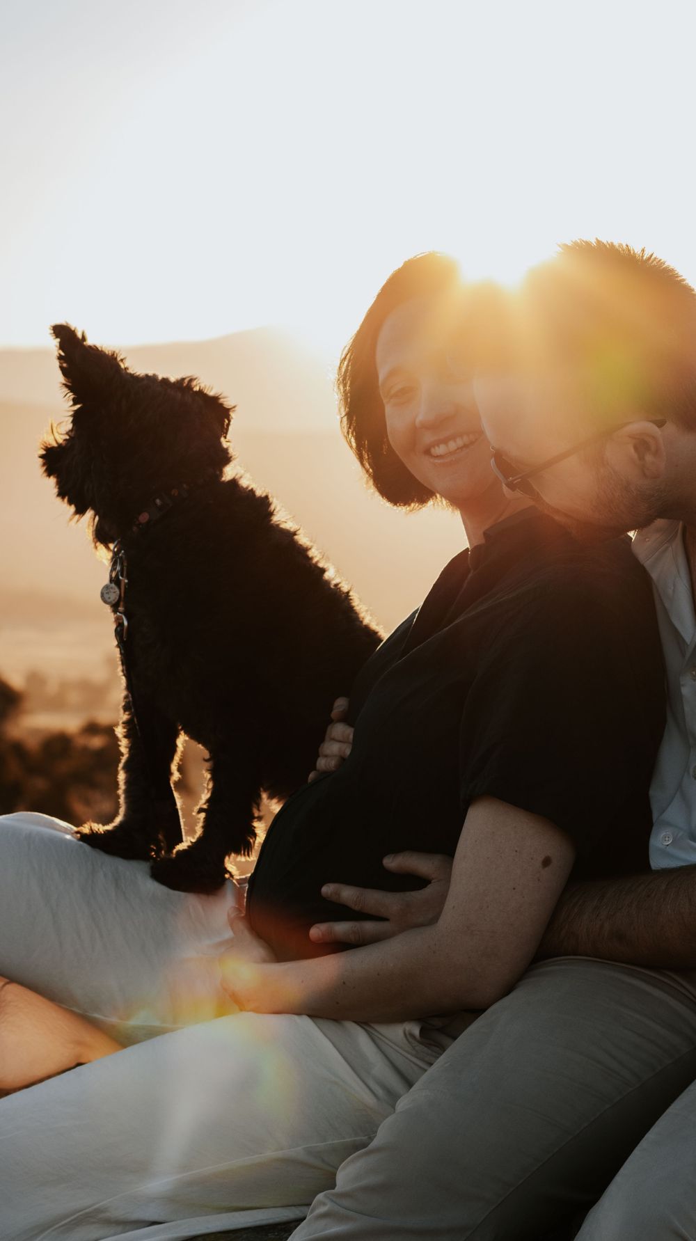 Couple sitting outdoors at sunset with a small black dog on pregnant woman's lap, golden backlight glowing warmly.