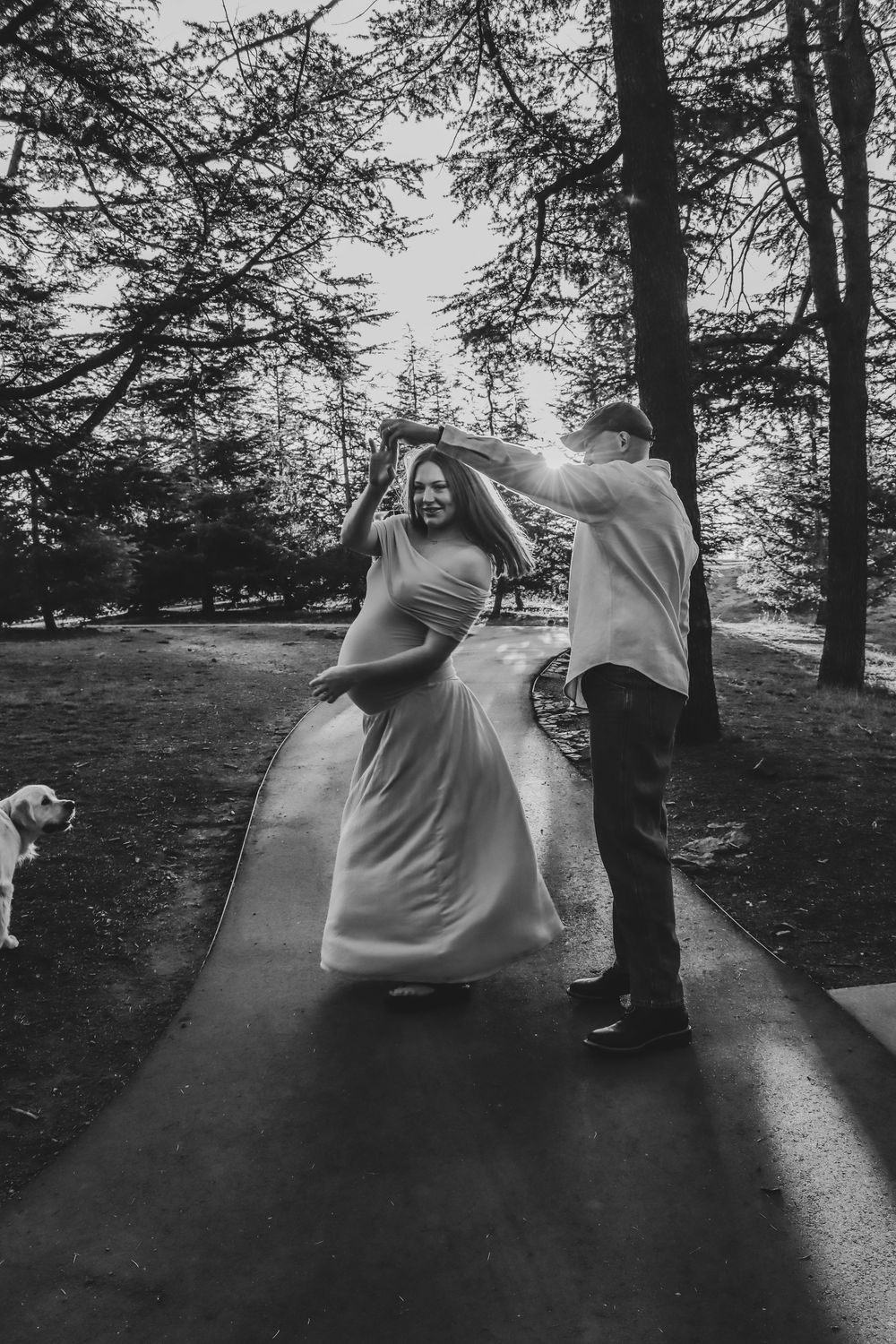 Black and white photo of a couple dancing on a path in a park, with a dog visible in the background.