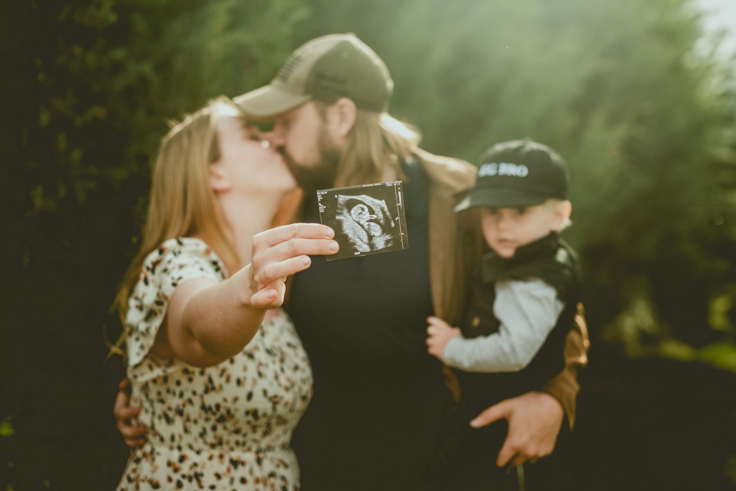 Couple kissing outdoors while holding ultrasound photo and toddler in pregnancy announcement portrait.