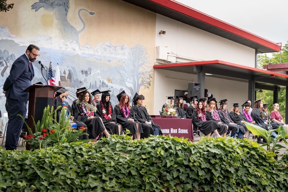 Speaker addresses graduates in caps and gowns at an outdoor high school commencement ceremony with mural backdrop.