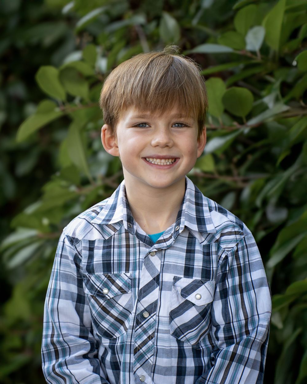 Smiling young boy with brown hair wearing a plaid shirt, posing outdoors with green foliage background.