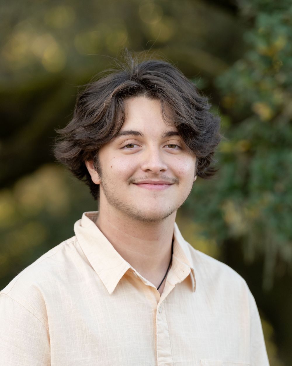 Young man with wavy dark hair smiling outdoors, wearing a light beige shirt with green trees in background.