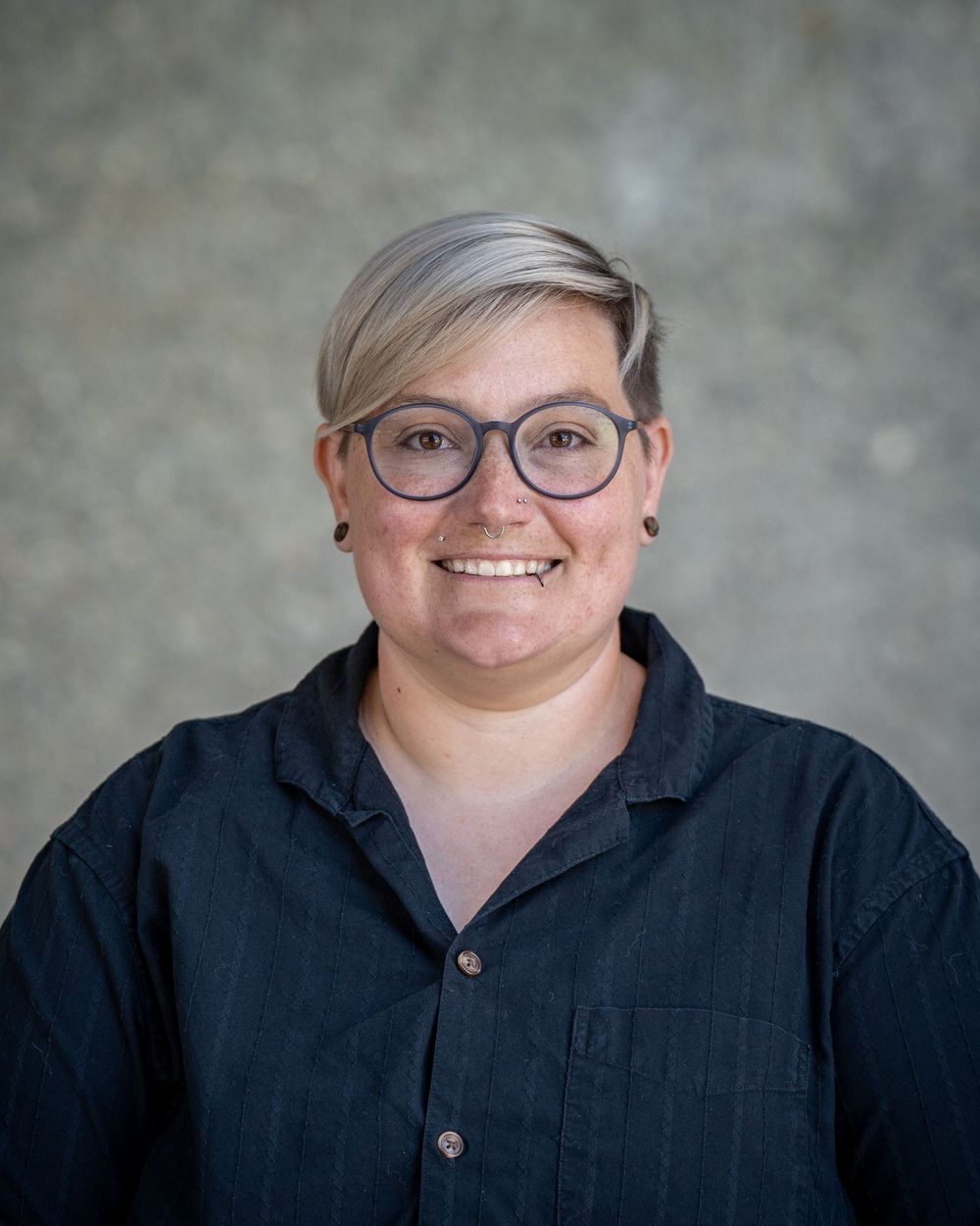 Smiling person with short gray hair and round glasses wearing a dark denim shirt against a gray background.