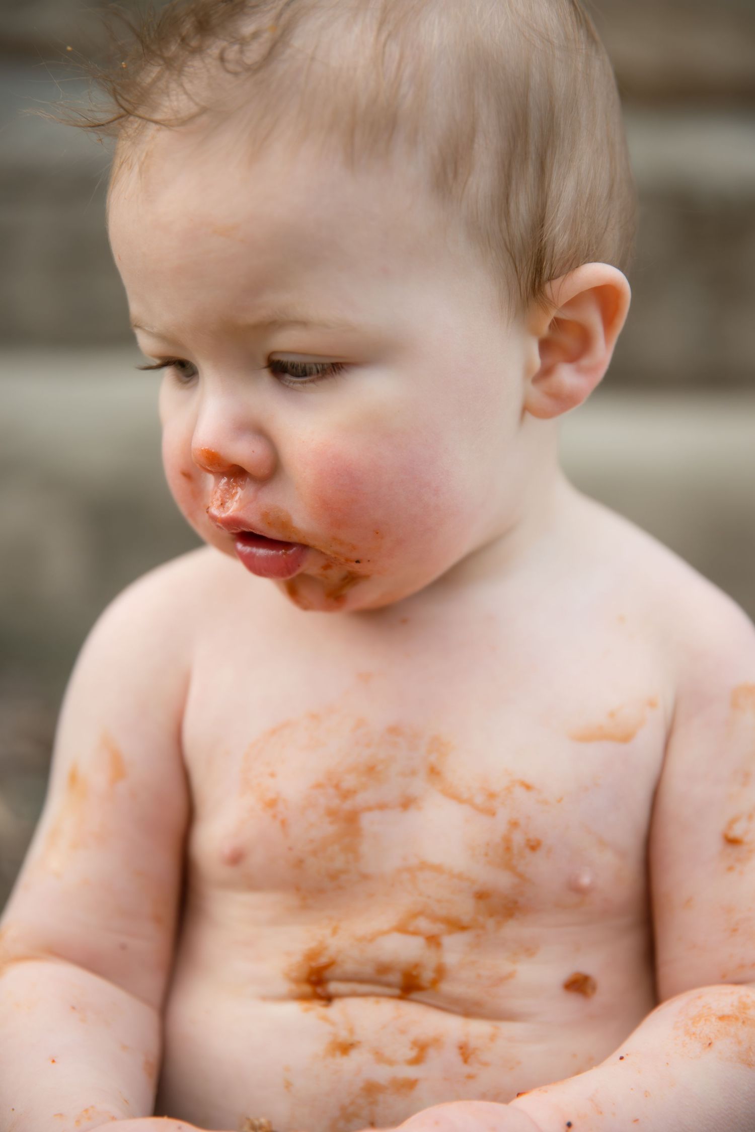 Messy baby covered in tomato sauce smiling, shirtless toddler with food on face and chest.