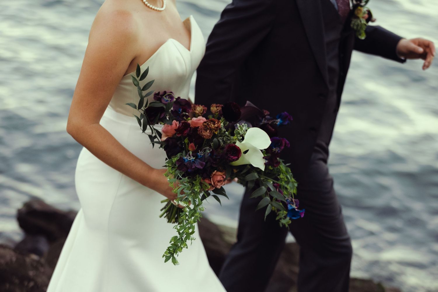 Bride in white gown holding dark floral bouquet with groom in suit near rocky waterfront.