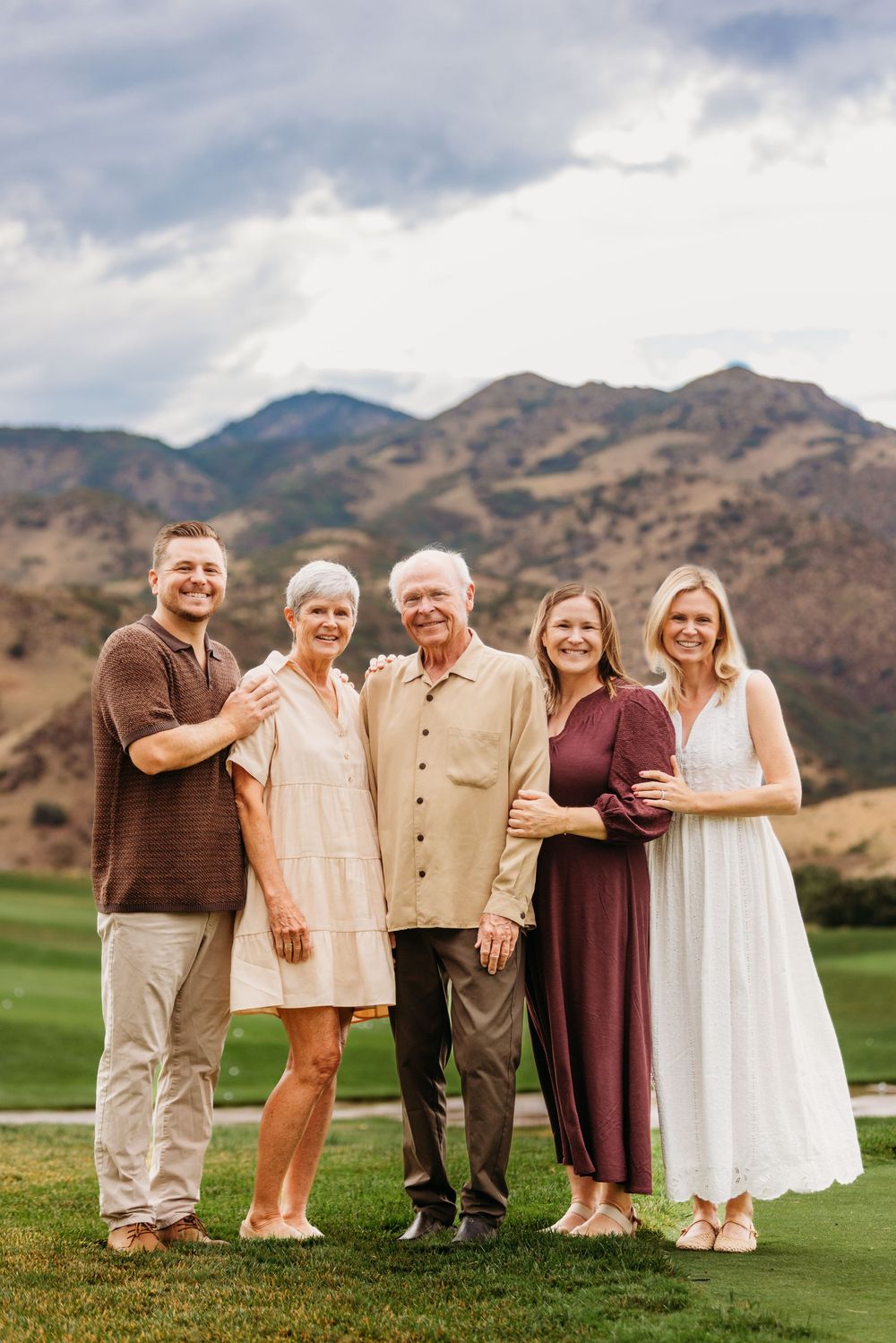 Family of five posing outdoors with mountain backdrop on green lawn under cloudy skies.
