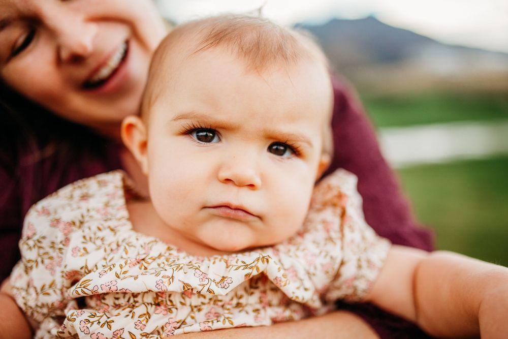 A baby in a floral outfit gazes at the camera while being held by a smiling woman outdoors.