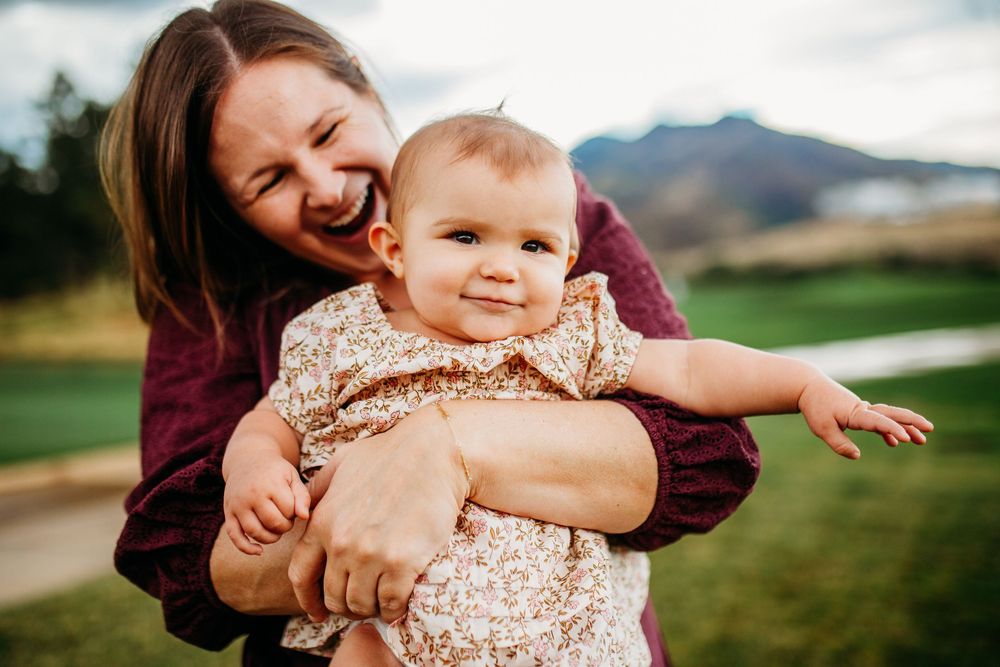 A laughing mother holds her smiling baby outdoors with mountains in the background.