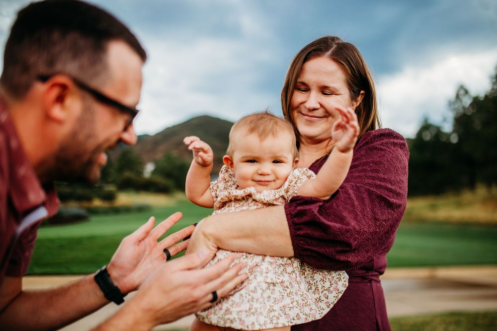 A smiling mother holds her happy baby girl while the father reaches out during a family photo session outdoors.