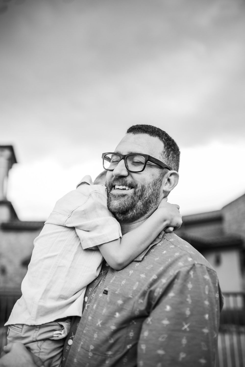 A bearded father wearing glasses laughs joyfully as his young child hugs him from behind in a black and white portrait.