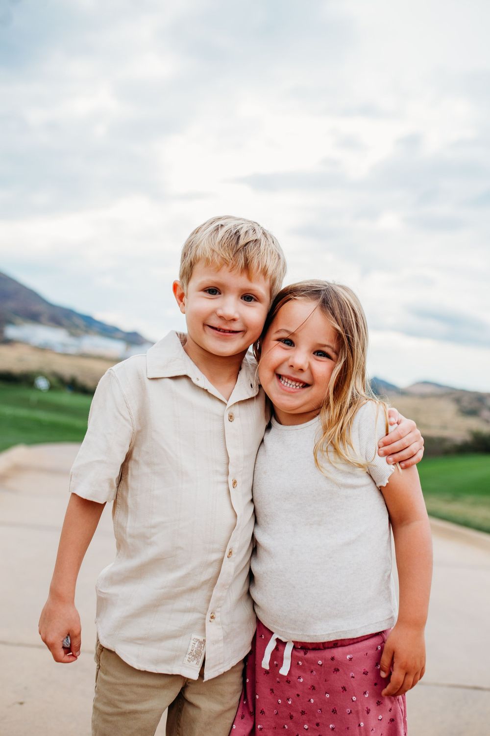 Two young siblings smiling and hugging outdoors with mountains and cloudy sky in the background.