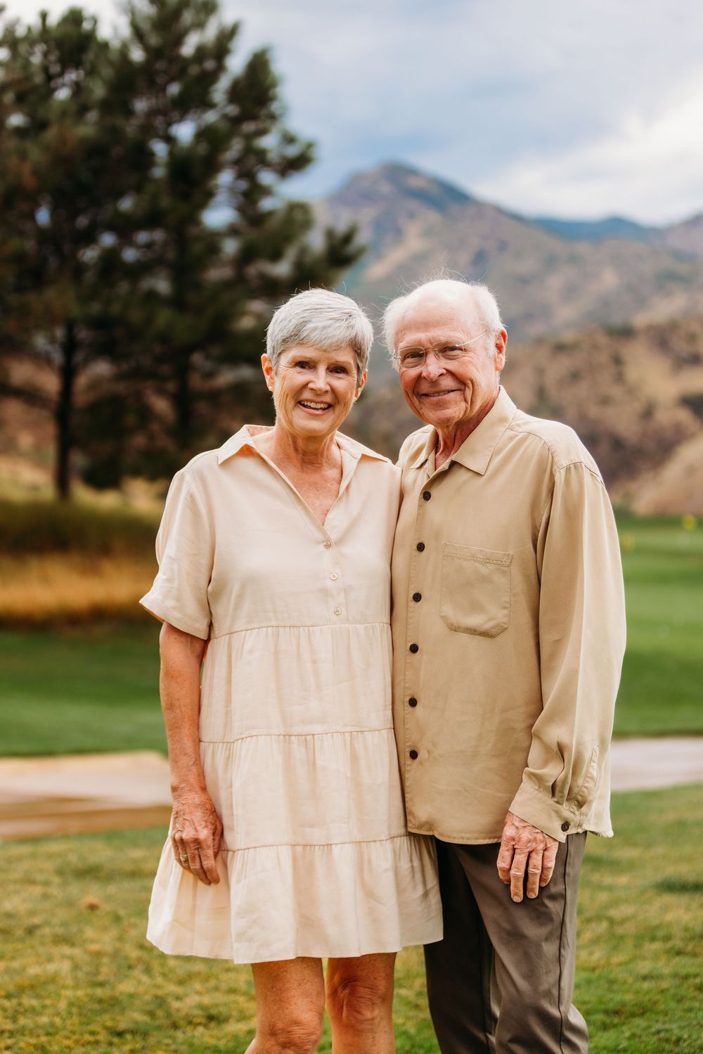 Smiling senior couple posing outdoors on a golf course with mountains and trees in the background.