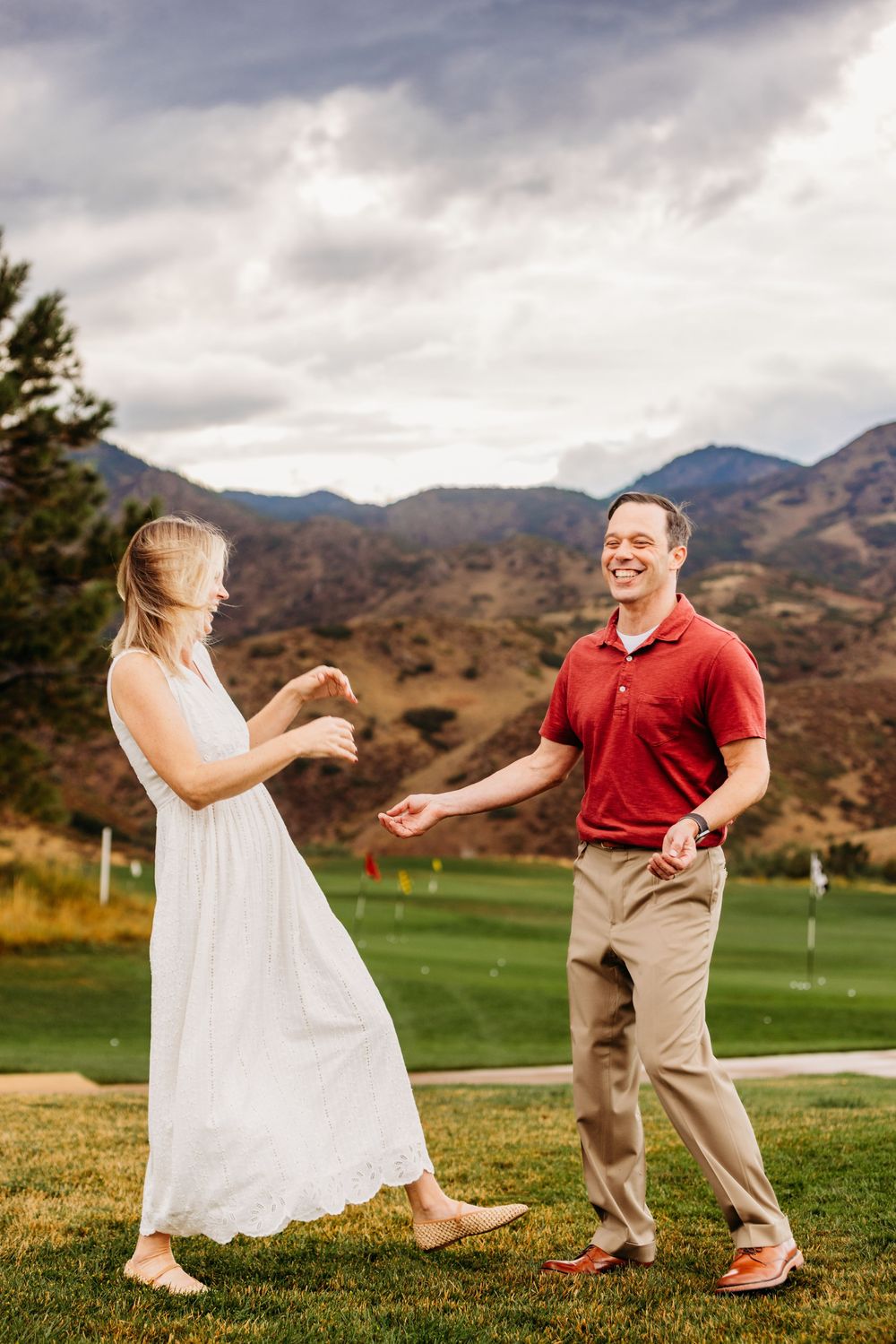 A smiling couple dances playfully on a golf course with mountains and dramatic cloudy sky behind them.