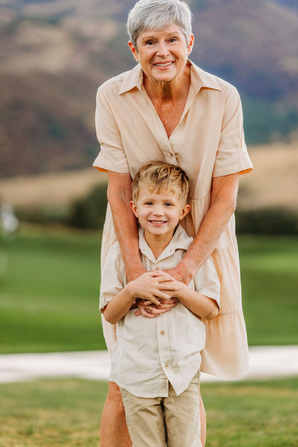 Elderly woman in beige dress hugging young blonde boy outdoors on green lawn with mountain backdrop.