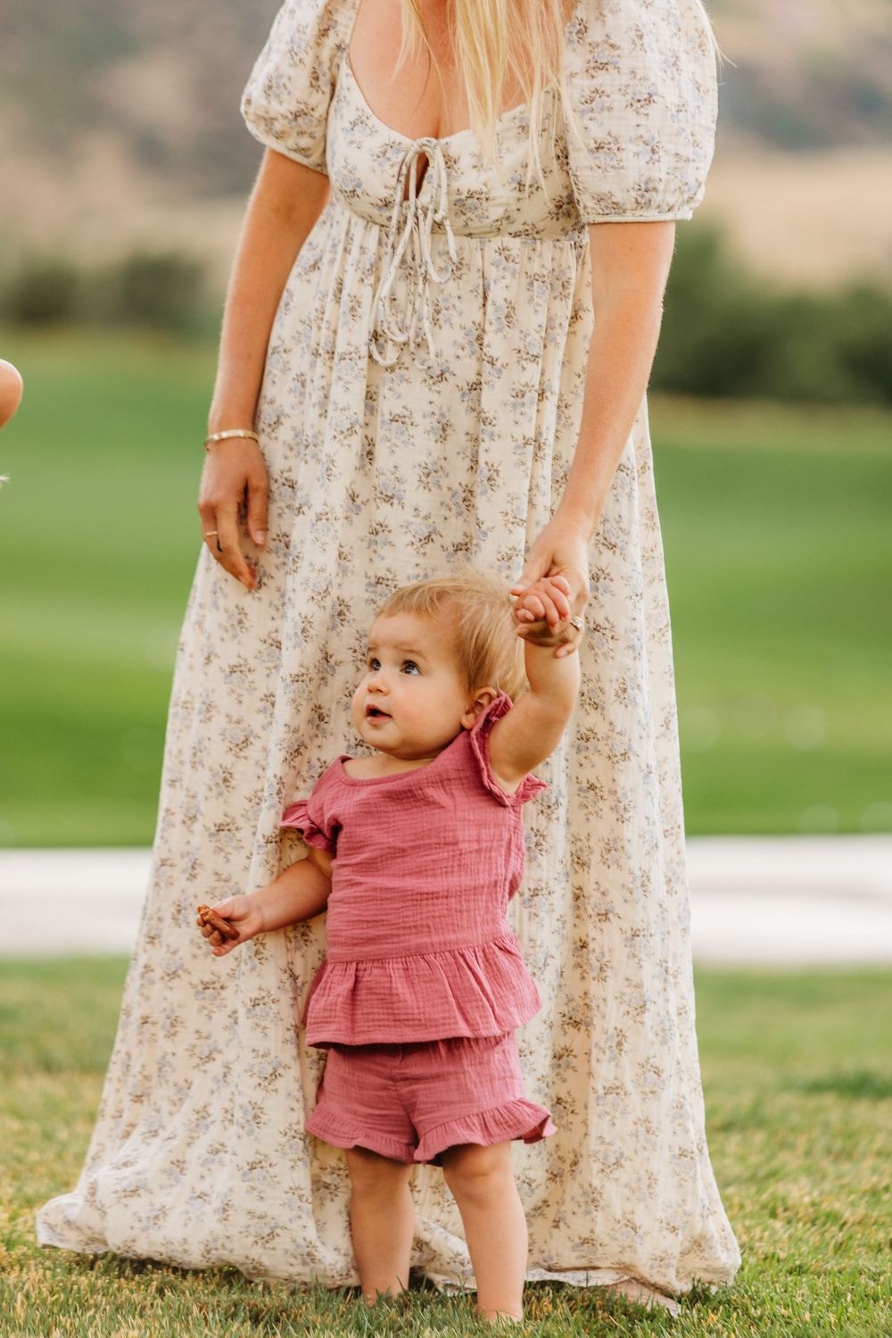 A baby girl in pink outfit holds her mother's hand while learning to walk on green grass.