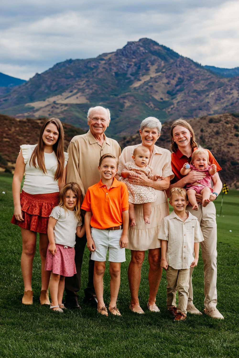 Multi-generational family portrait outdoors on green grass with mountain backdrop, including grandparents, teens, and young children.