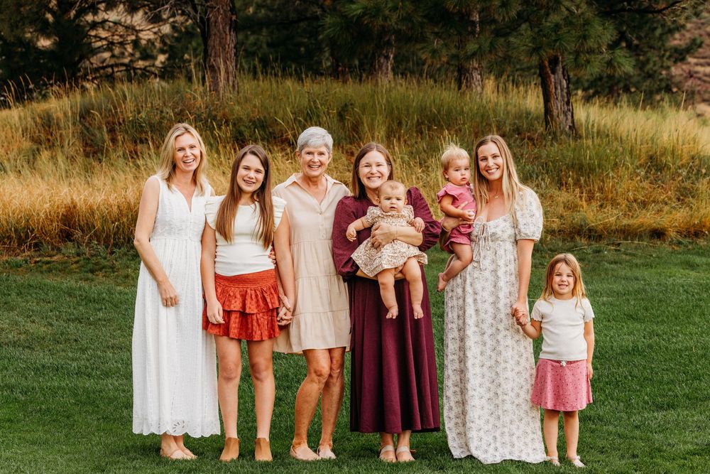 A multigenerational family of women and girls smiling together outdoors on a green lawn surrounded by trees.