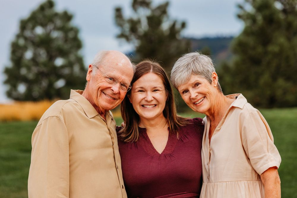 Three smiling adults pose together outdoors on a green field with trees and mountains in the background.