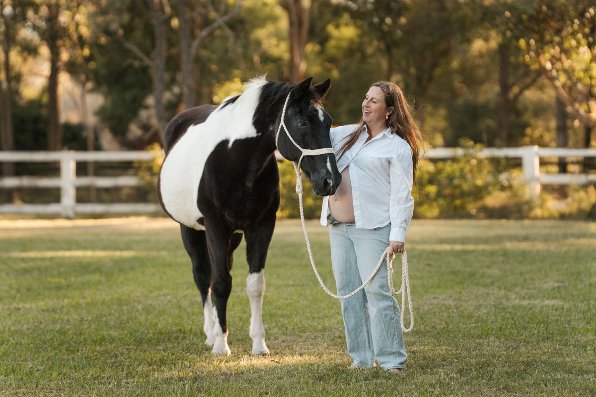 Woman in light blue jeans, leading a black & white paint horse on a green lawn near white fence, maternity photoshoot with a horse in sydney