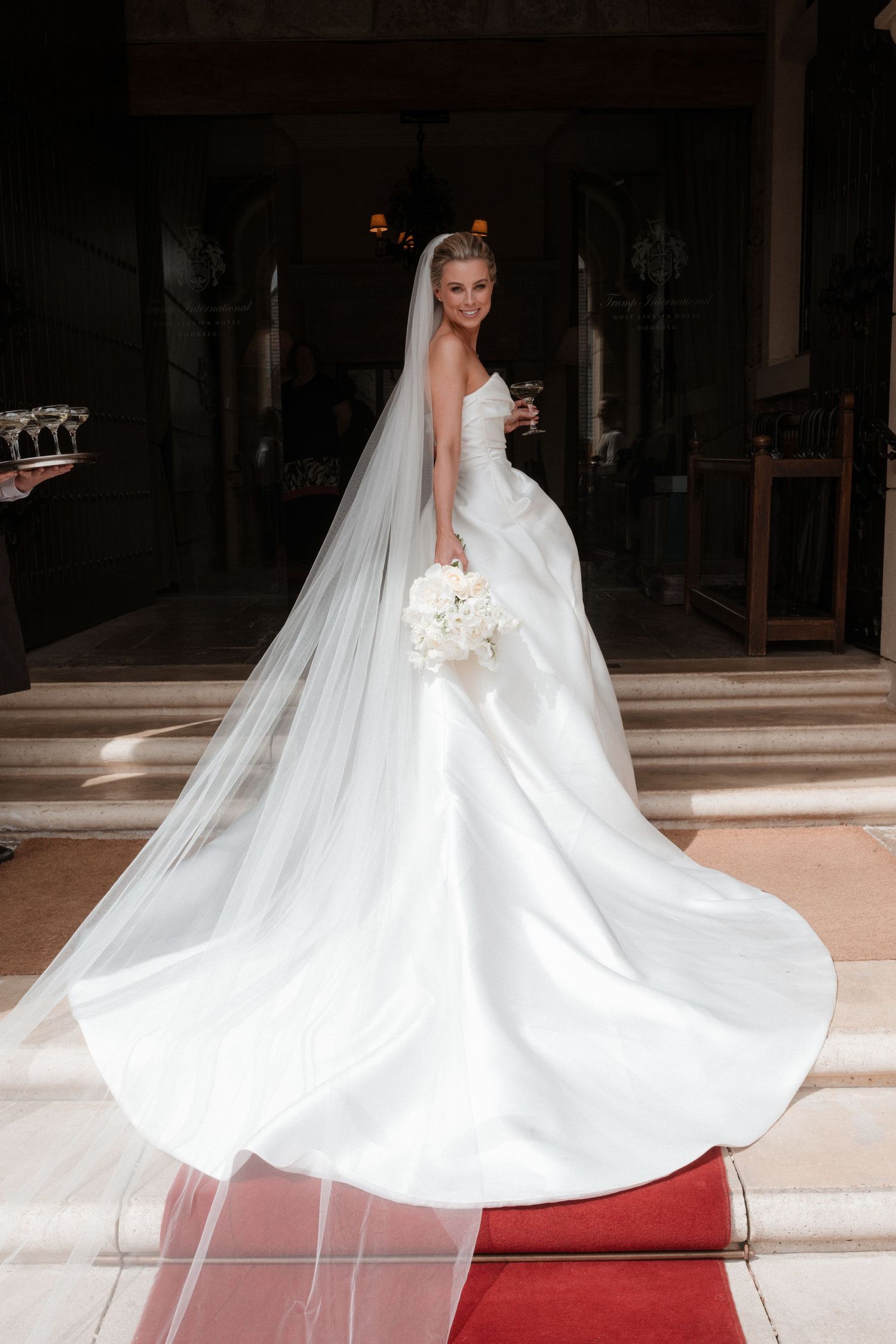 Elegant bride in a stunning white ball gown with long veil holding bouquet on red carpet steps.