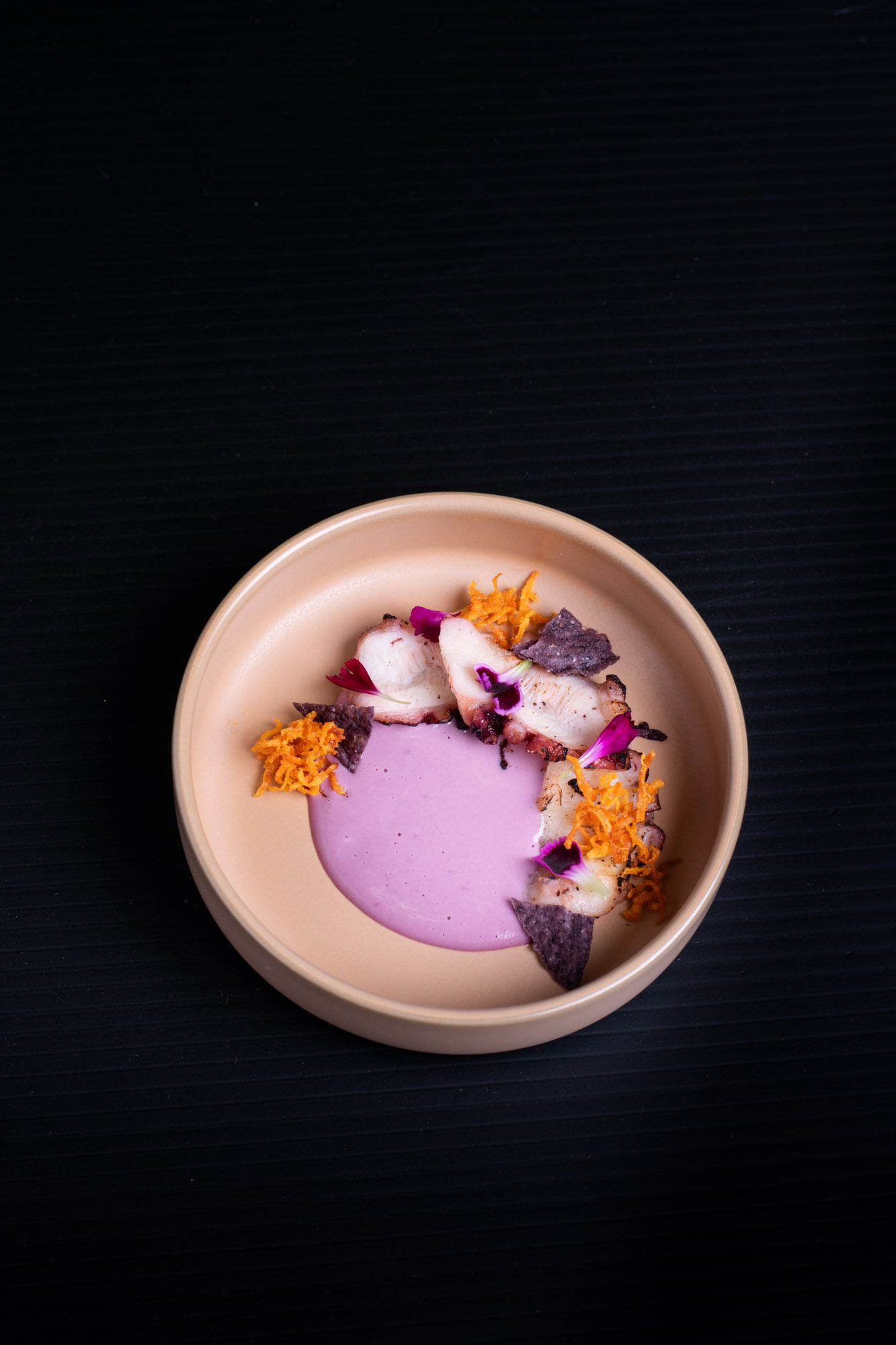 Elegant pink soup in a beige bowl, garnished with edible flowers, orange zest, and purple herbs on a dark background.