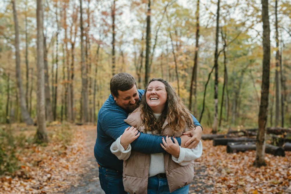 Couple laughing and embracing on a leaf-covered forest path surrounded by autumn foliage.