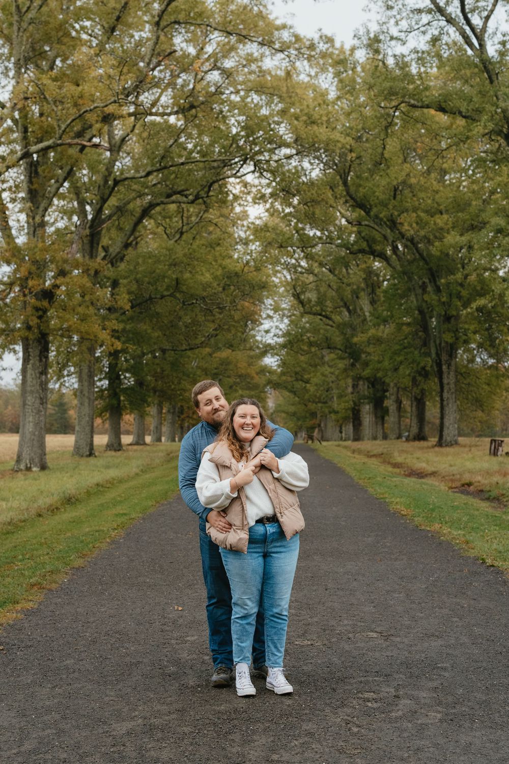 A smiling couple embraces on a tree-lined path in autumn, wearing casual jeans and jackets during an outdoor engagement photo.