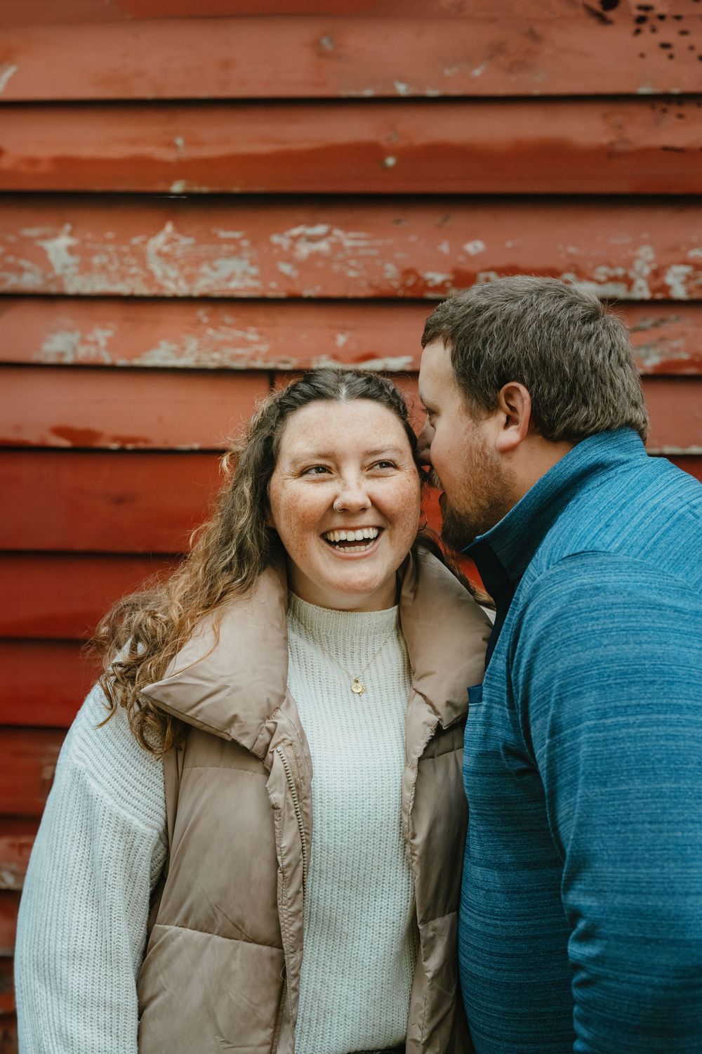 A smiling woman in a puffer vest laughs as a man whispers in her ear near a rustic red barn wall.