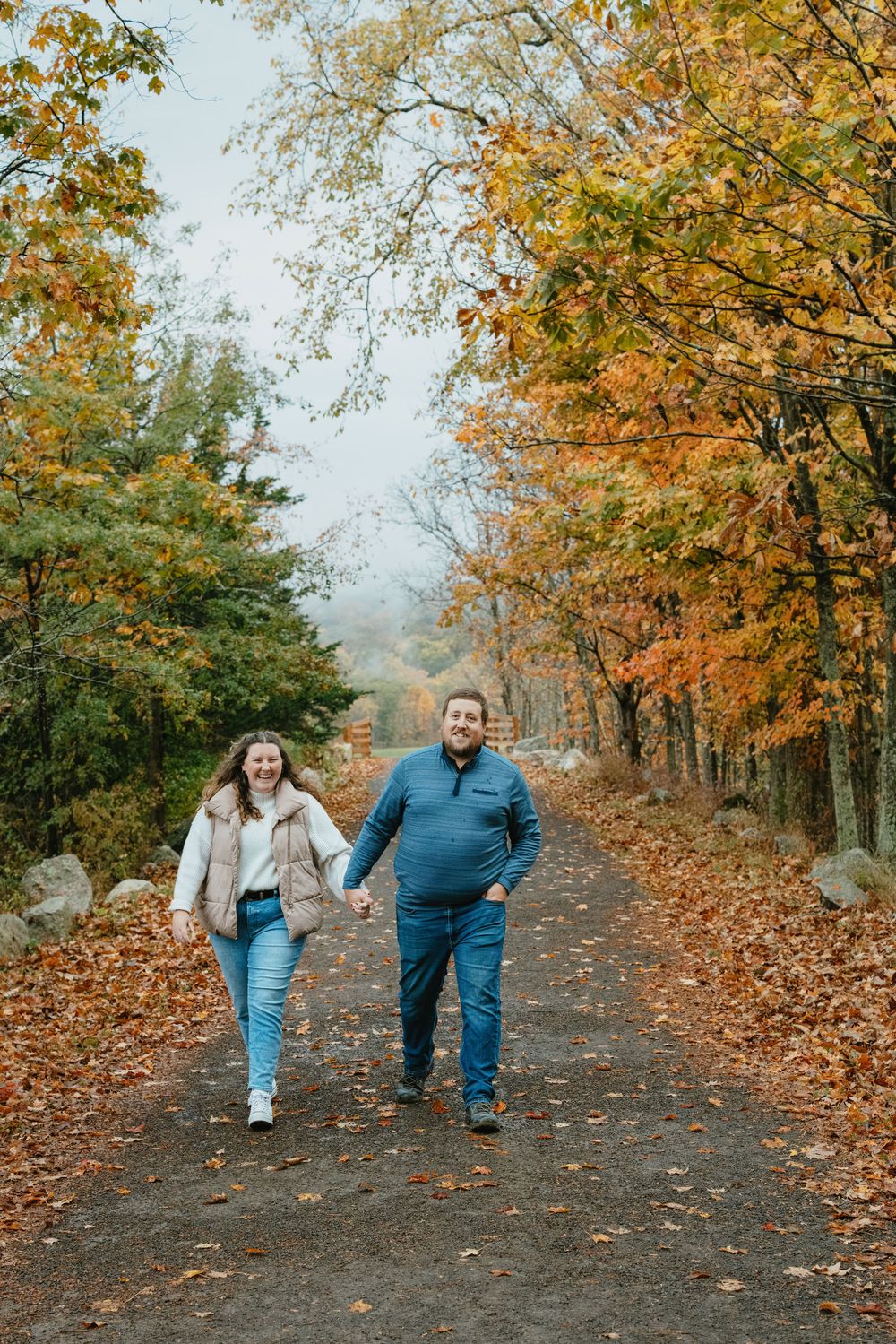 Couple holding hands and walking on a leaf-covered path surrounded by colorful autumn foliage.