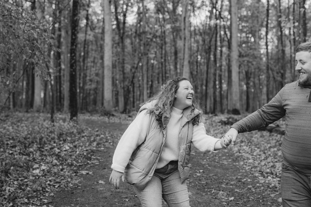 Black and white photo of a laughing couple holding hands and walking through an autumn forest.