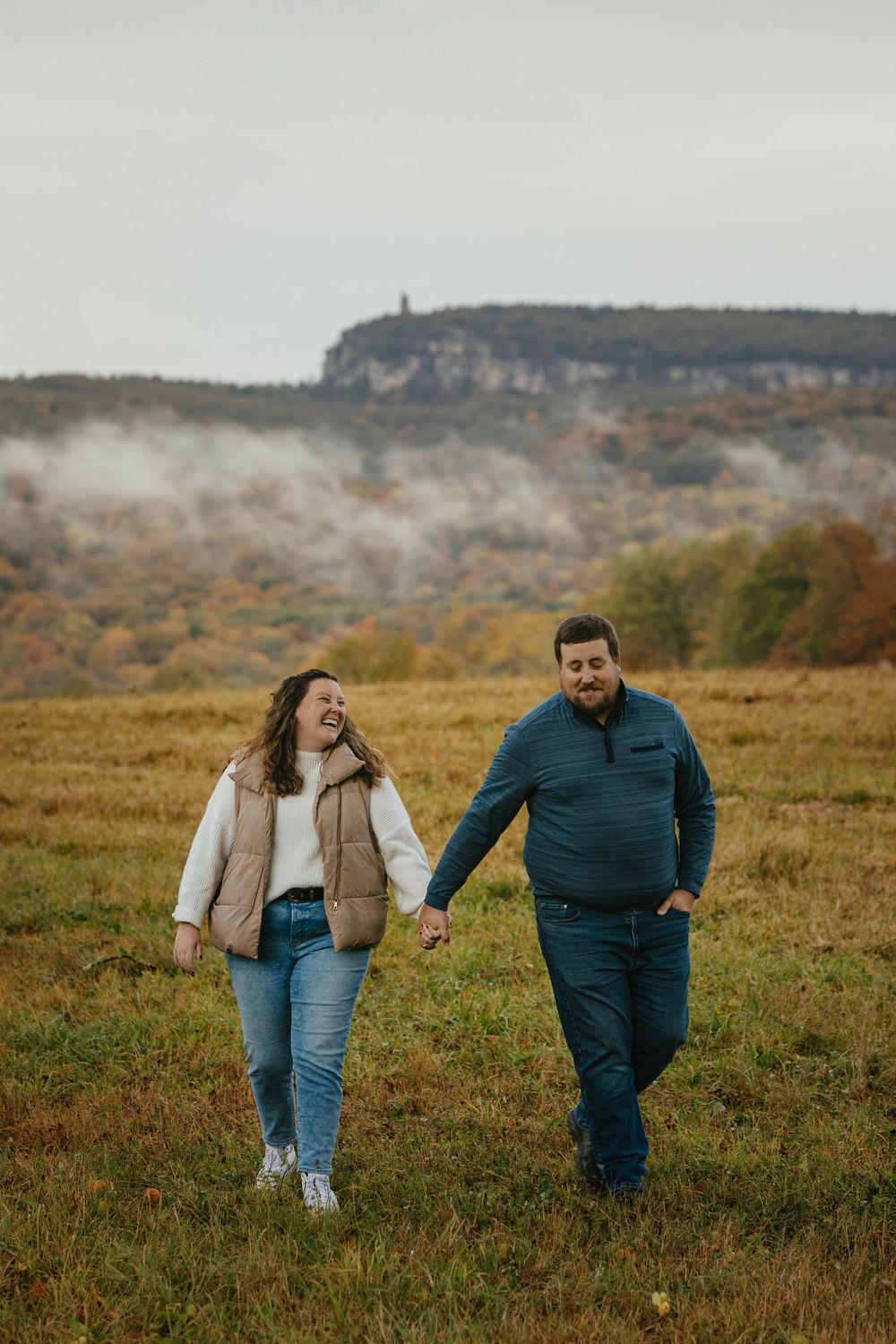 A couple holds hands and laughs while walking through an autumn field with misty cliffs in the background.
