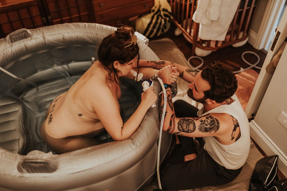 Tattooed couple sharing an intimate moment beside an inflatable birth pool during a home water birth.