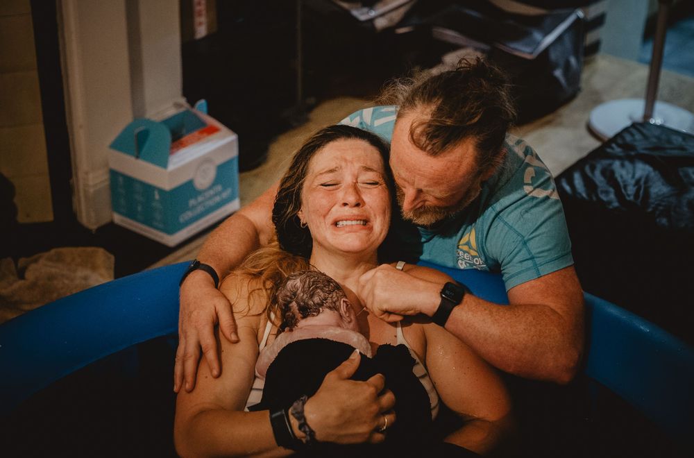 Emotional water birth moment with mother holding newborn, supported by partner in birthing pool at home.
