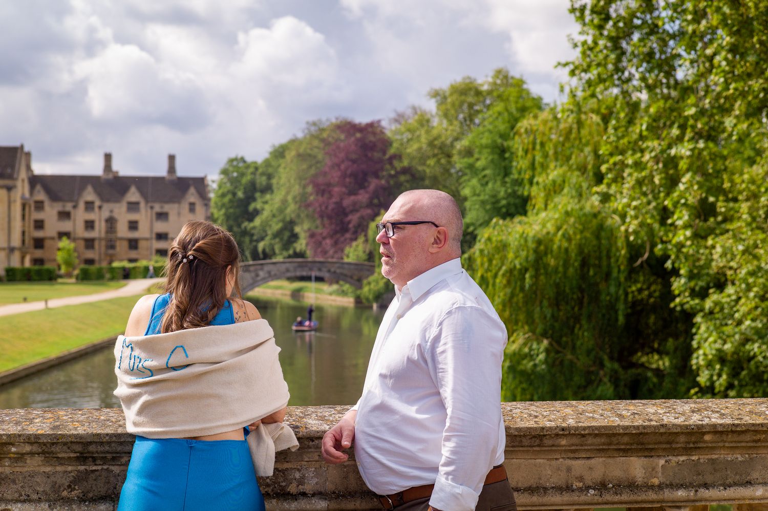 A man and woman stand by a stone bridge overlooking a scenic river with punting boats and lush green trees.