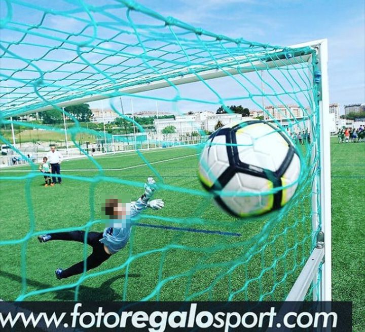 Un balón de fútbol entra en la portería mientras el portero se lanza para atajarlo durante un partido en un campo de césped verde.