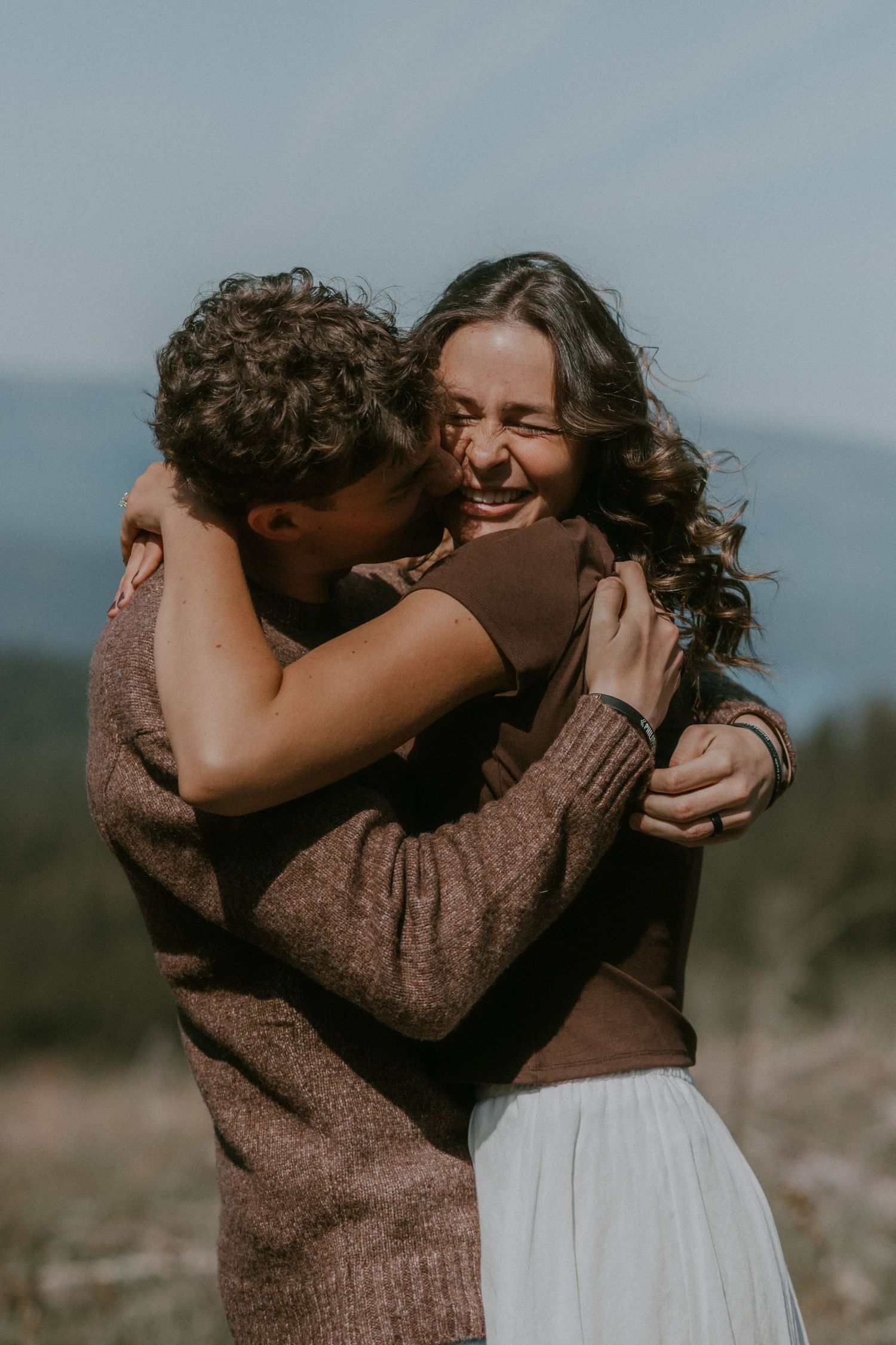 Couple embracing outdoors, woman laughing joyfully as man kisses her cheek, wearing cozy sweaters with mountain backdrop.