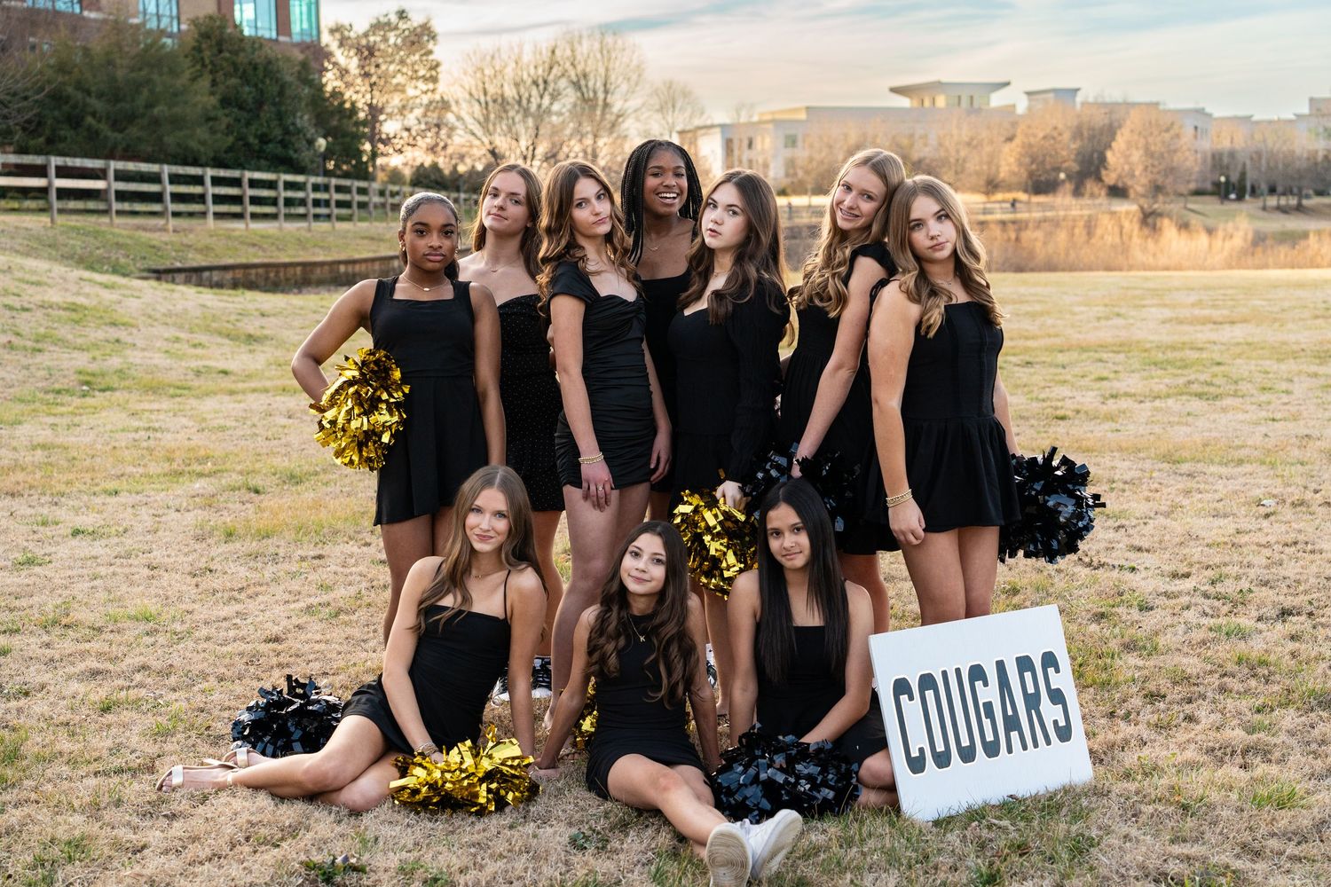 Group of cheerleaders in black and white uniforms posing outdoors for a team photo with gold pom-poms.