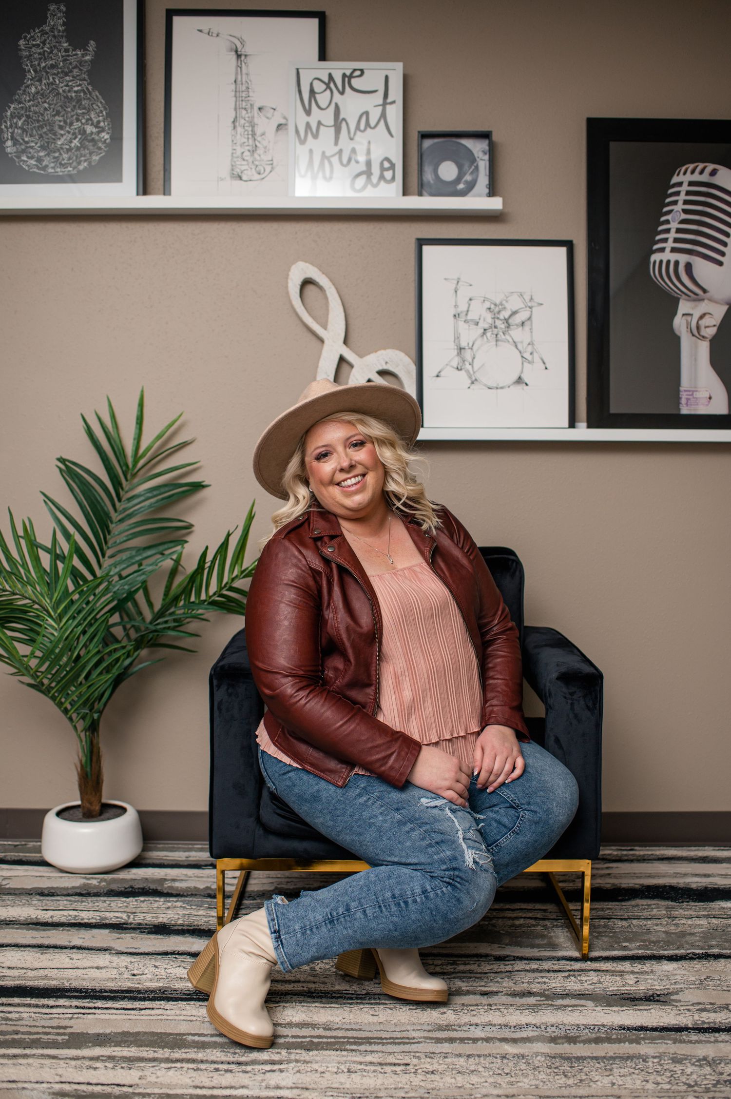 Smiling blonde woman in burgundy leather jacket, hat, and jeans seated in velvet chair with music-themed decor backdrop.