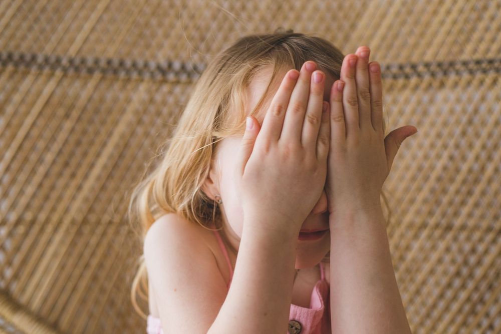 Young blonde girl covering her face with both hands, wearing a pink outfit.