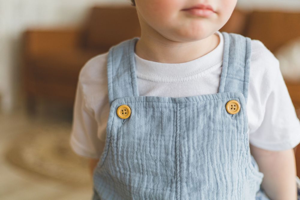 Cute toddler wearing a light blue linen overalls with wooden buttons over a white t-shirt.