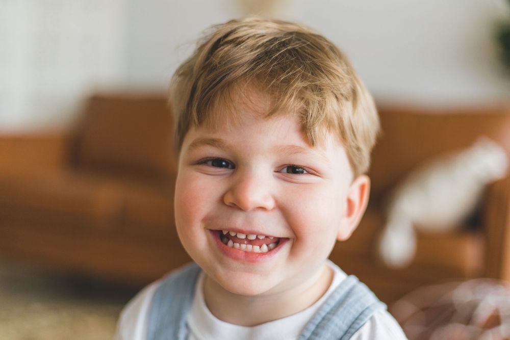 Happy toddler boy with blonde hair laughing and smiling brightly in a warm cozy living room setting.