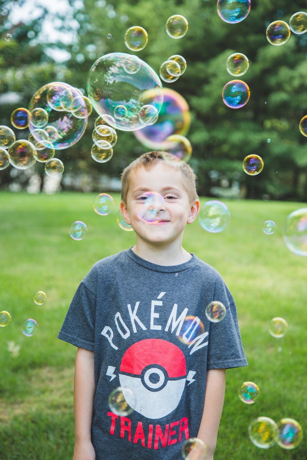 Young boy in a Pokemon Trainer t-shirt smiling outdoors surrounded by colorful floating soap bubbles in a green park.