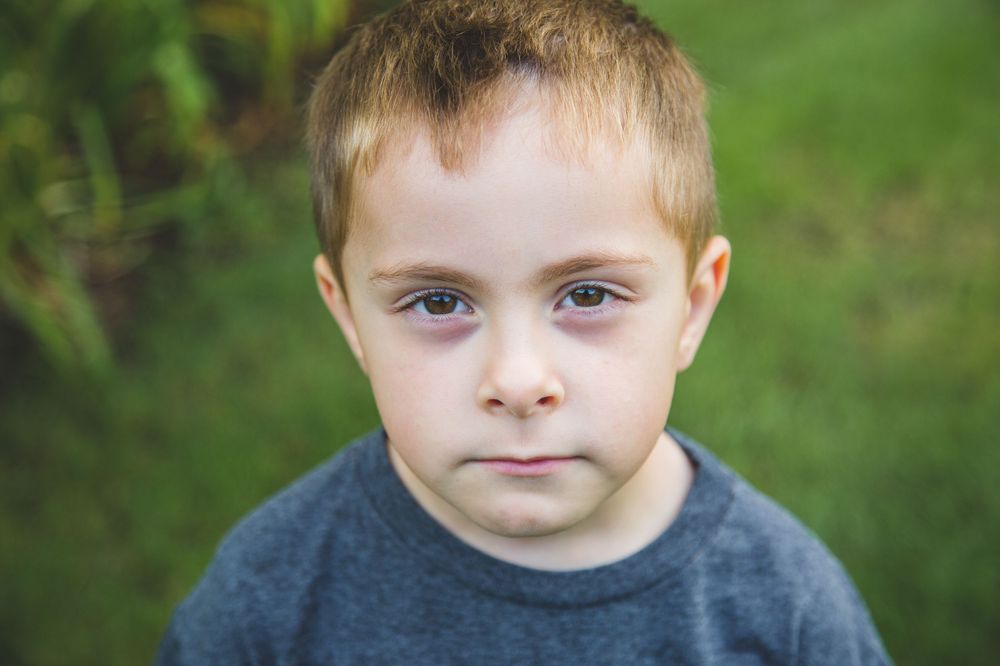 A young boy with blonde hair wearing a gray shirt looks seriously at the camera outdoors with green grass behind him.