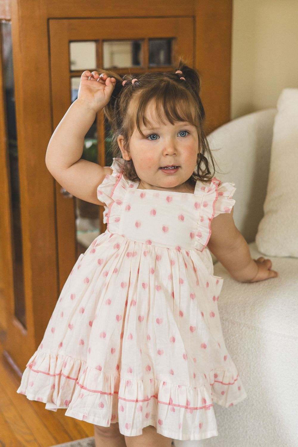 Toddler girl in white pink polka dot ruffle dress standing indoors touching her hair pigtails.