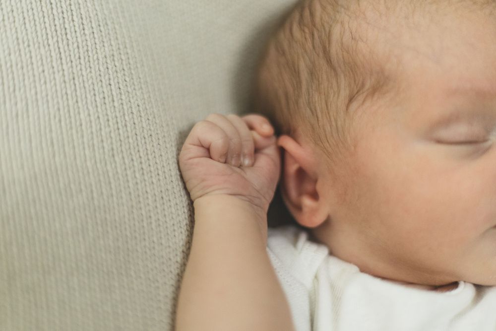 Sleeping newborn baby with tiny clenched fist resting against a soft beige surface, peaceful close-up portrait.