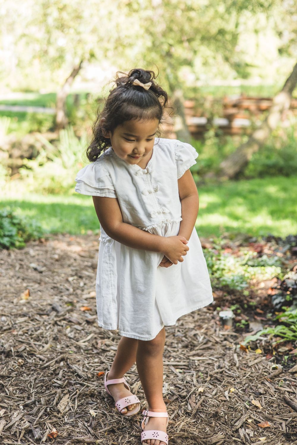 Young girl in white dress and pink sandals standing in a garden, looking down with hands clasped.