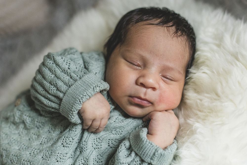 Peaceful newborn baby sleeping in sage green knit outfit nestled on a soft white fluffy blanket.