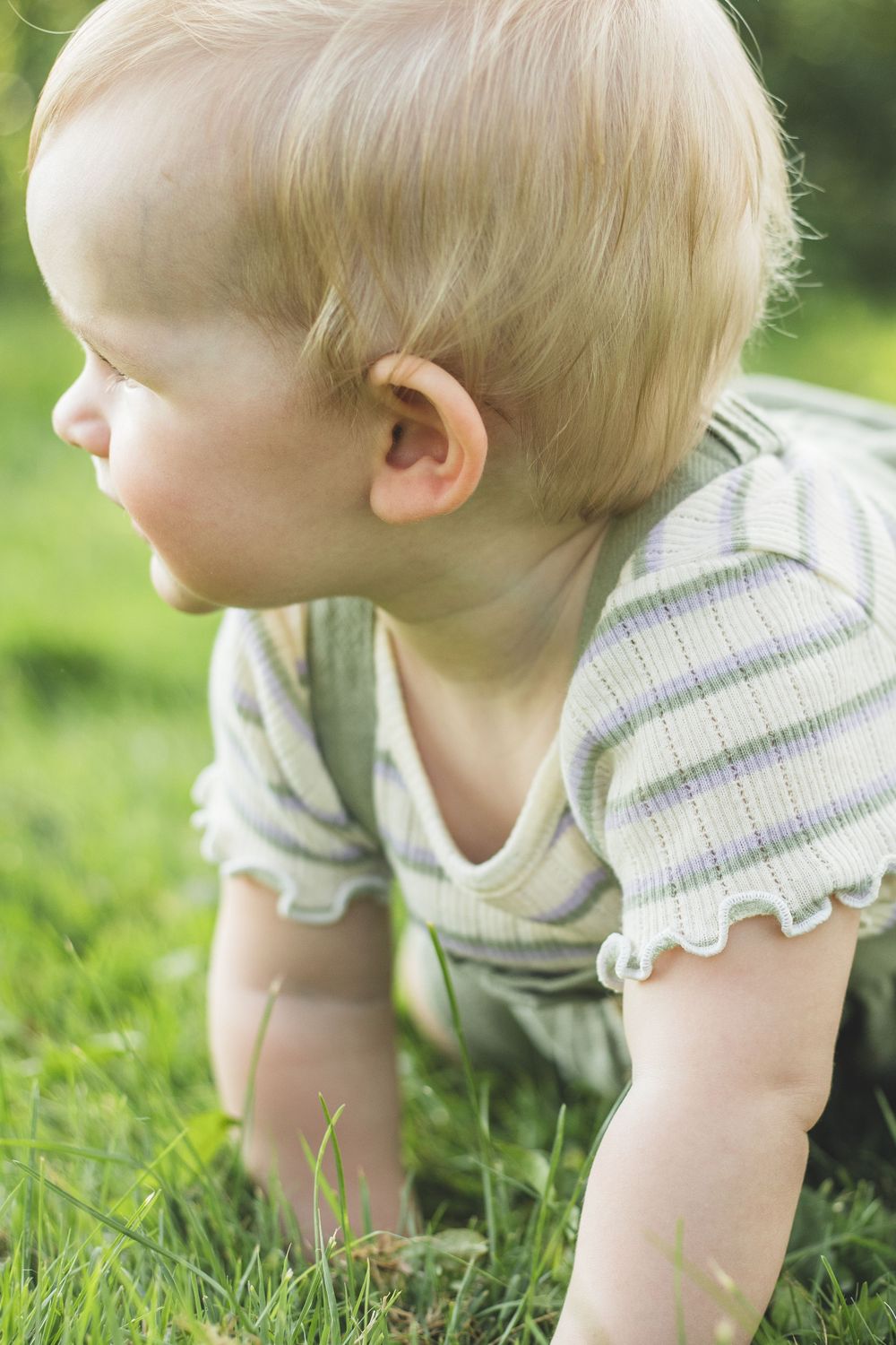 Blonde toddler crawling on green grass outdoors, wearing a striped onesie on a sunny day.